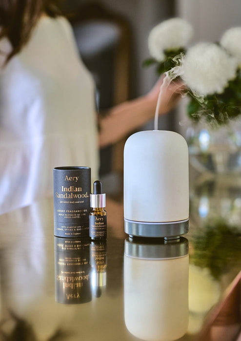 indian sandalwood fragrance oil displayed in home setting next to electric diffuser with person arranging flowers in the bacground